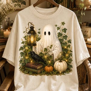 May include: An off-white t-shirt featuring a graphic print of a white ghost holding a lit lantern, surrounded by green ivy, a black crow perched on two stacked books, and two pumpkins. The text PURE GRAPH is visible on the left and right sides.