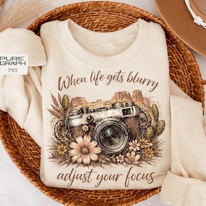 May include: Cream-colored sweatshirt featuring a vintage camera design with the text "When life gets blurry, adjust your focus." The graphic includes desert landscape elements and floral accents. The sweatshirt is folded in a woven basket.