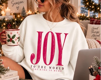 May include: A white crewneck sweatshirt featuring the word JOY in large, textured pink letters, with the text TO THE WORLD THE KING IS HERE printed below in smaller black font. The garment has long sleeves and a classic, relaxed fit design.