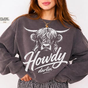 May include: Charcoal grey sweatshirt with a white Highland cow graphic and the text "Howdy darlin'". The design is printed on a crew neck, long-sleeved top. The style is casual and evokes a Western aesthetic, suitable for everyday wear.