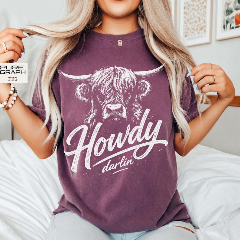 May include: A purple t-shirt with a white Highland cow illustration and the text "Howdy darlin'". The cow has long horns and shaggy fur. The text is in a cursive font. The shirt is a solid colour.