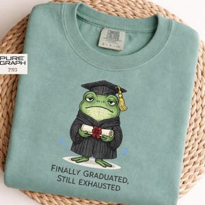 Puede incluir: Una sudadera verde agua con una rana de dibujos animados con birrete y toga, sosteniendo un diploma. El texto "FINALLY GRADUATED, STILL EXHAUSTED" está impreso debajo. La sudadera tiene cuello redondo y una etiqueta Comfort Colors.