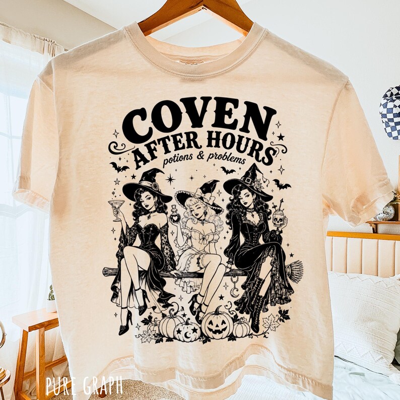 Puede incluir: Una camiseta de color crema con un estampado gr&aacute;fico negro de tres brujas sentadas en una escoba. El texto dice COVEN AFTER HOURS y potions & problems. El dise&ntilde;o incluye estrellas, murci&eacute;lagos, calabazas y hojas en una ilustraci&oacute;n detallada.