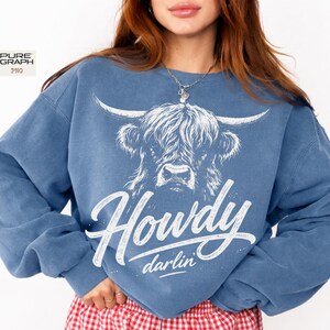 May include: A blue sweatshirt with a white graphic of a Highland cow and the text "Howdy darlin'". The sweatshirt has a crew neck and long sleeves. The design is in a script font.