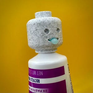 May include: A white plastic tube with a purple label that reads "2X FASTER WHITENING", "RECYCLE", and "NET WT 3.5 OZ". The tube has a white Lego minifigure head on top with a blue tongue sticking out.
