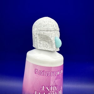Pode incluir: Um tubo de plástico branco com uma tampa de garrafa em forma de capacete mandaloriano cinza e branco. O tubo é etiquetado com "NET WT 3.8 OZ (107 g)" e "MINT RADIANT".