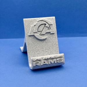 May include: A speckled light grey phone stand featuring the Los Angeles Rams logo embossed at the top and the text LOS ANGELES RAMS embossed on the front ledge. The stand has a slanted back support and a horizontal base for holding a device.