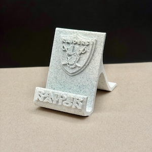 Puede incluir: Un soporte para teléfono impreso en 3D de color blanco moteado con el logotipo del escudo de los Raiders en relieve en el panel posterior y el texto RAIDERS NATION en relieve en el borde frontal. El soporte tiene un diseño inclinado.