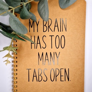 May include: A brown spiral notebook with the text "My brain has too many tabs open." printed in black.
