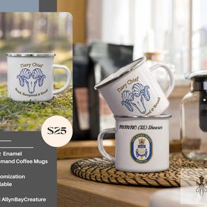 May include: Two white enamel mugs with blue and gold designs. The top mug has the text "Navy Chief" and a ram's head with the text "Tried, Tempered & True". The bottom mug has the text "mmnc (SS) Shearer" and a navy seal emblem. The mugs are sitting on a woven placemat.