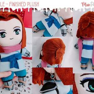 May include: A plush toy with brown hair, a blue and white outfit, and a pink scarf. The toy is wearing a blue bow around its waist. The toy is sitting on a table with a red and white pattern.