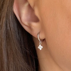 May include: A silver hoop earring with a small, four-petal flower charm. The charm is adorned with small, clear gemstones. The earring is shown on a person's ear, with their skin and hair visible in the background.