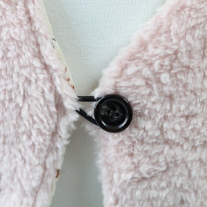 May include: A close-up of a pink fluffy fabric with a black button and elastic band.