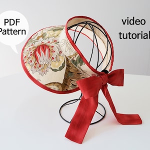 May include: A floral patterned sun hat with a red trim and a red bow. The hat is on a black stand. The text "PDF Pattern" and "video tutorial" is visible in the image.