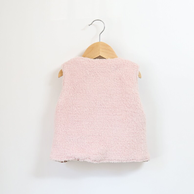 May include: A pink fleece vest with no sleeves hanging on a wooden hanger.