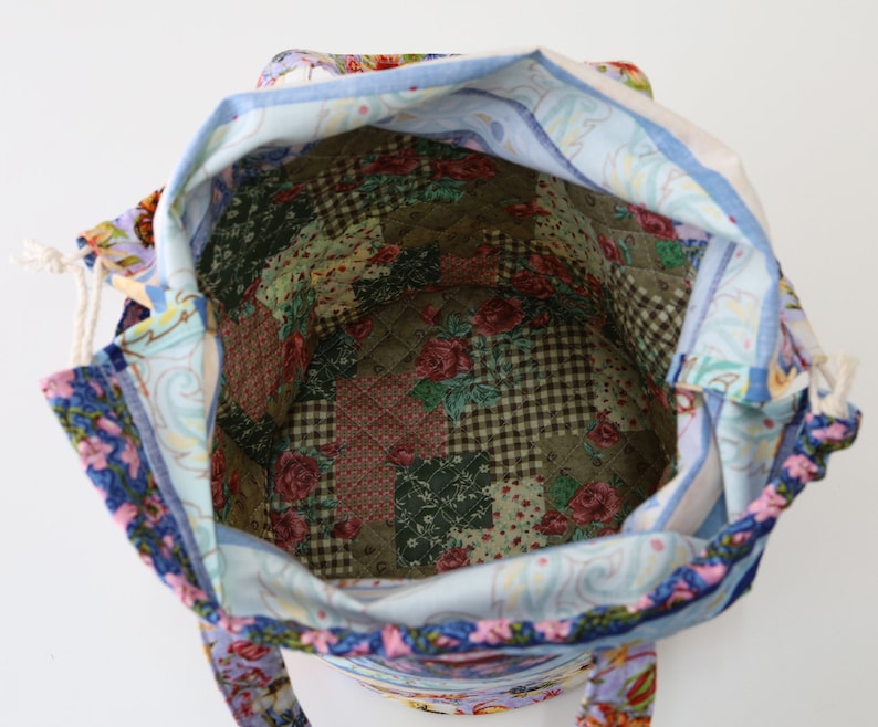 K&ouml;nnte beinhalten: Eine blaue und wei&szlig;e Stofftasche mit Blumendruck und Kordelzug. Das Innere der Tasche ist mit einem Patchwork-Stoff ausgekleidet, der verschiedene Blumen- und Karomuster aufweist.