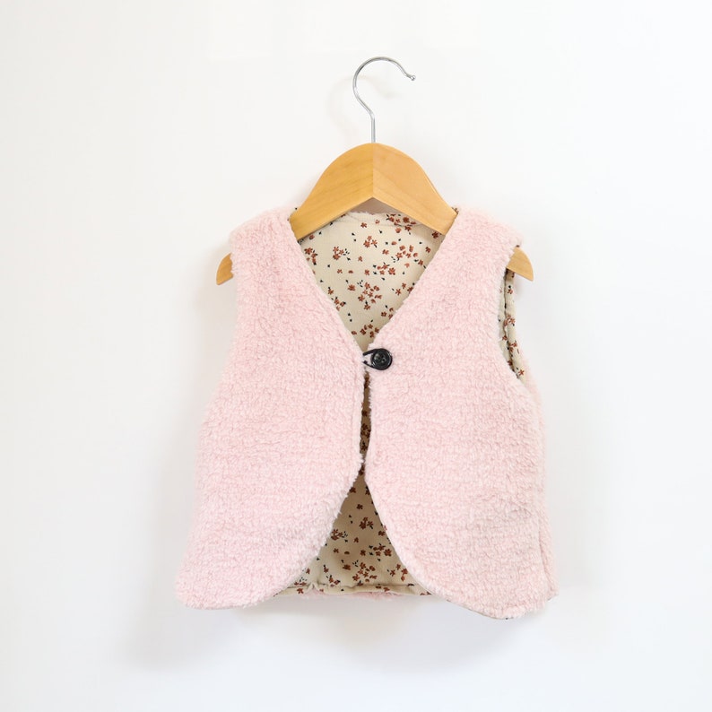 May include: A pink fleece vest with a floral print lining. The vest has a single button closure and is hanging on a wooden hanger.