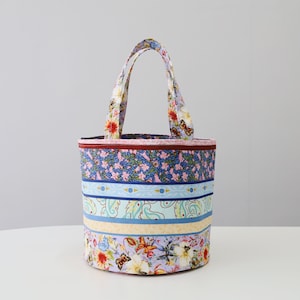 K&ouml;nnte beinhalten: Eine bunte Stofftasche mit Blumenmuster und blauen und gelben Streifen. Die Tasche hat zwei Henkel und eine zylindrische Form.
