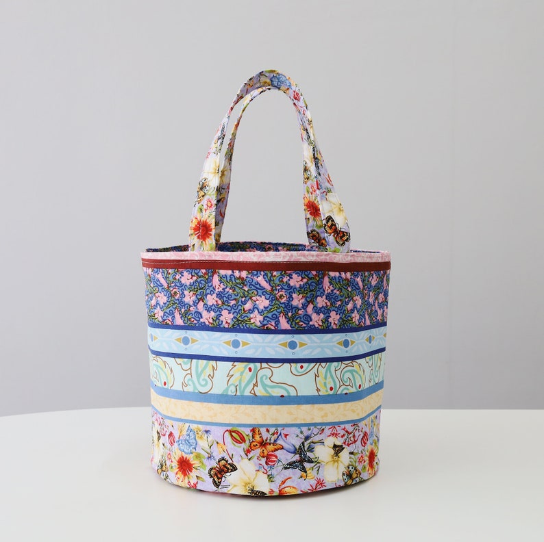 K&ouml;nnte beinhalten: Eine bunte Stofftasche mit Blumenmuster und blauen und gelben Streifen. Die Tasche hat zwei Henkel und eine zylindrische Form.