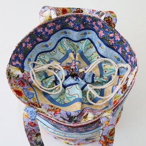 K&ouml;nnte beinhalten: Eine farbenfrohe Tragetasche mit Kordelzug und Blumenmuster. Die Tasche hat ein blaues, gr&uuml;nes und pinkfarbenes Blumenmuster auf wei&szlig;em Hintergrund. Die Tasche ist aus Stoff und hat einen wei&szlig;en Kordelzug.