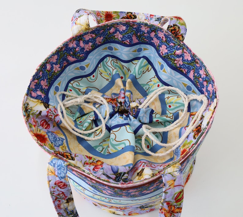 K&ouml;nnte beinhalten: Eine farbenfrohe Tragetasche mit Kordelzug und Blumenmuster. Die Tasche hat ein blaues, gr&uuml;nes und pinkfarbenes Blumenmuster auf wei&szlig;em Hintergrund. Die Tasche ist aus Stoff und hat einen wei&szlig;en Kordelzug.