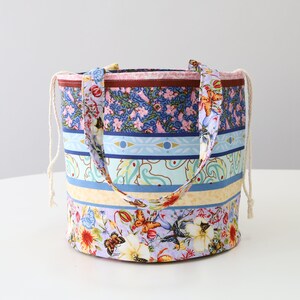 K&ouml;nnte beinhalten: Eine bunte gestreifte Stofftasche mit Kordelzug. Die Tasche hat ein Blumenmuster mit Schmetterlingen und Paisley-Mustern in Blaut&ouml;nen, Gelb, Rosa und Gr&uuml;n.