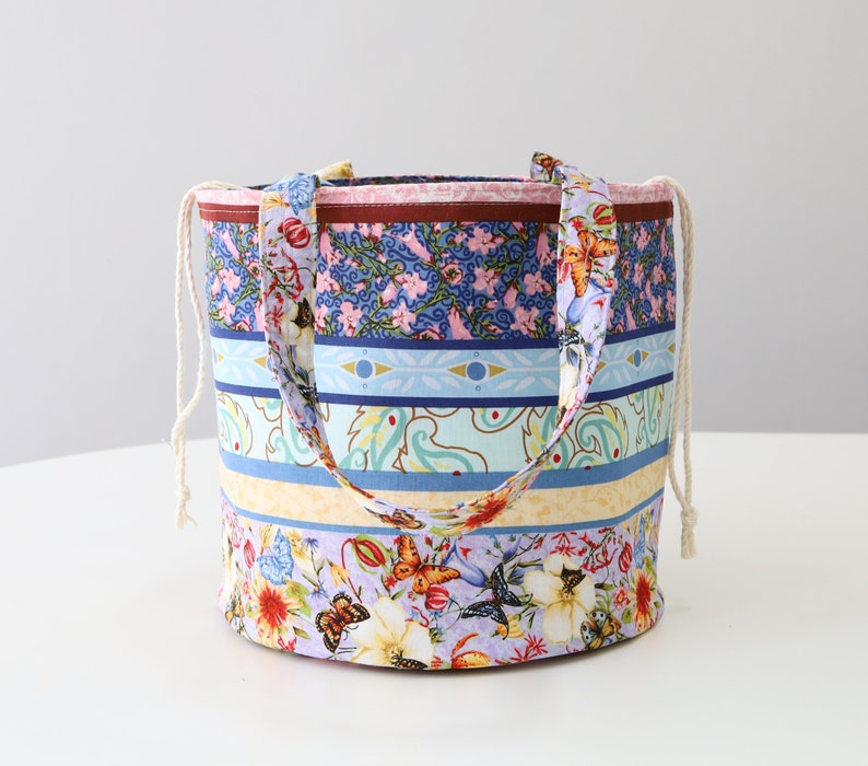 K&ouml;nnte beinhalten: Eine bunte gestreifte Stofftasche mit Kordelzug. Die Tasche hat ein Blumenmuster mit Schmetterlingen und Paisley-Mustern in Blaut&ouml;nen, Gelb, Rosa und Gr&uuml;n.