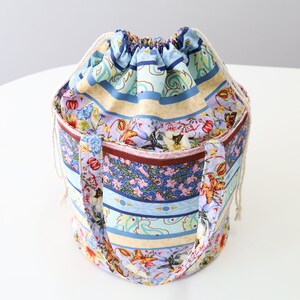 K&ouml;nnte beinhalten: Eine bunte Kordeltasche mit floralem und geometrischem Muster. Die Tasche hat eine hellblaue, gelbe, rosa und braune Farbgebung. Die Tasche hat zwei Henkel und einen Kordelverschluss.