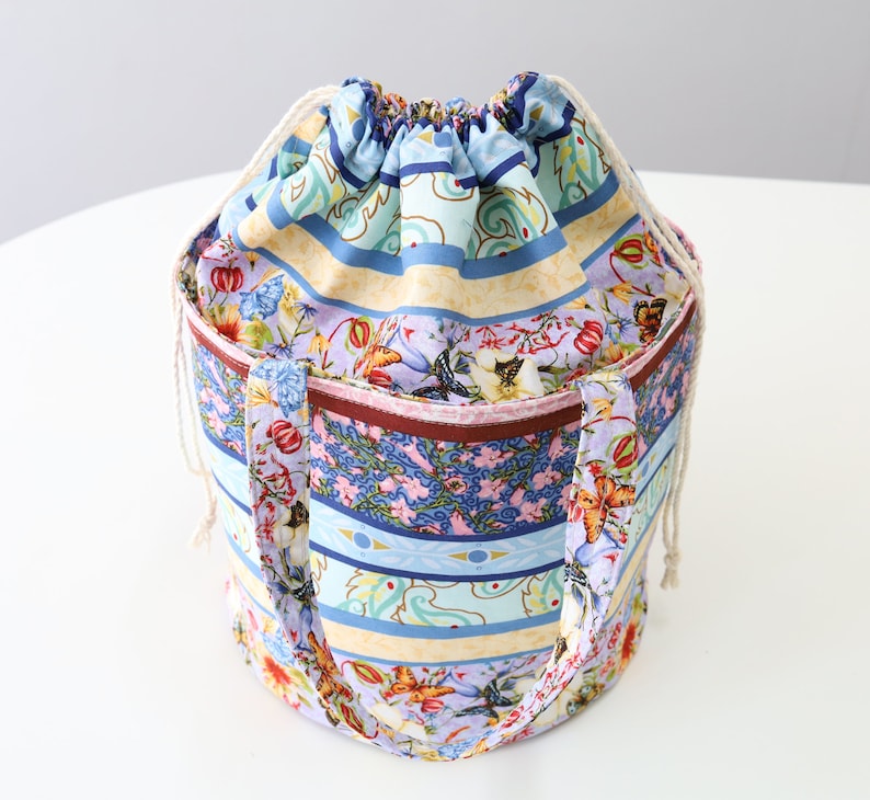 K&ouml;nnte beinhalten: Eine bunte Kordeltasche mit floralem und geometrischem Muster. Die Tasche hat eine hellblaue, gelbe, rosa und braune Farbgebung. Die Tasche hat zwei Henkel und einen Kordelverschluss.