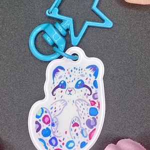 Peut inclure: Un porte-clés blanc et bleu avec un léopard des neiges de dessin animé avec des taches colorées. Le porte-clés est attaché à un anneau métallique en forme d'étoile bleue.