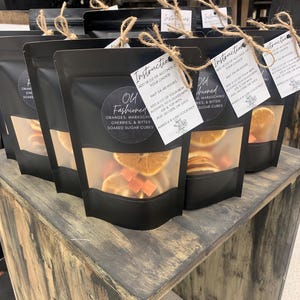 May include: Four black bags with clear windows filled with dried orange slices, maraschino cherries, and sugar cubes. Each bag has a white tag with the text "Old Fashioned" and instructions for making a cocktail.