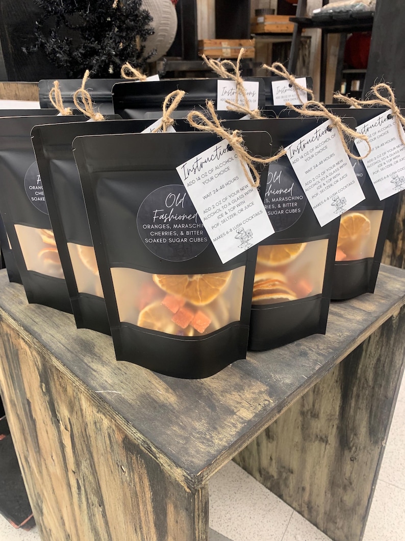 May include: Four black bags with clear windows filled with dried orange slices, maraschino cherries, and sugar cubes. Each bag has a white tag with the text "Old Fashioned" and instructions for making a cocktail.