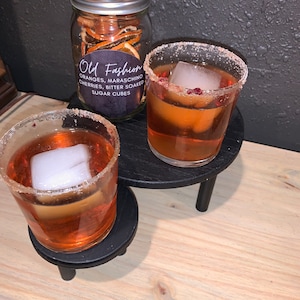 May include: Two clear glasses with orange liquid and ice cubes, rimmed with sugar. The glasses are on black coasters with three legs. A glass jar with orange peels and a label that reads "Old Fashioned Oranges, Maraschino Cherries, Bitter Soaked Sugar Cubes" is in the background.