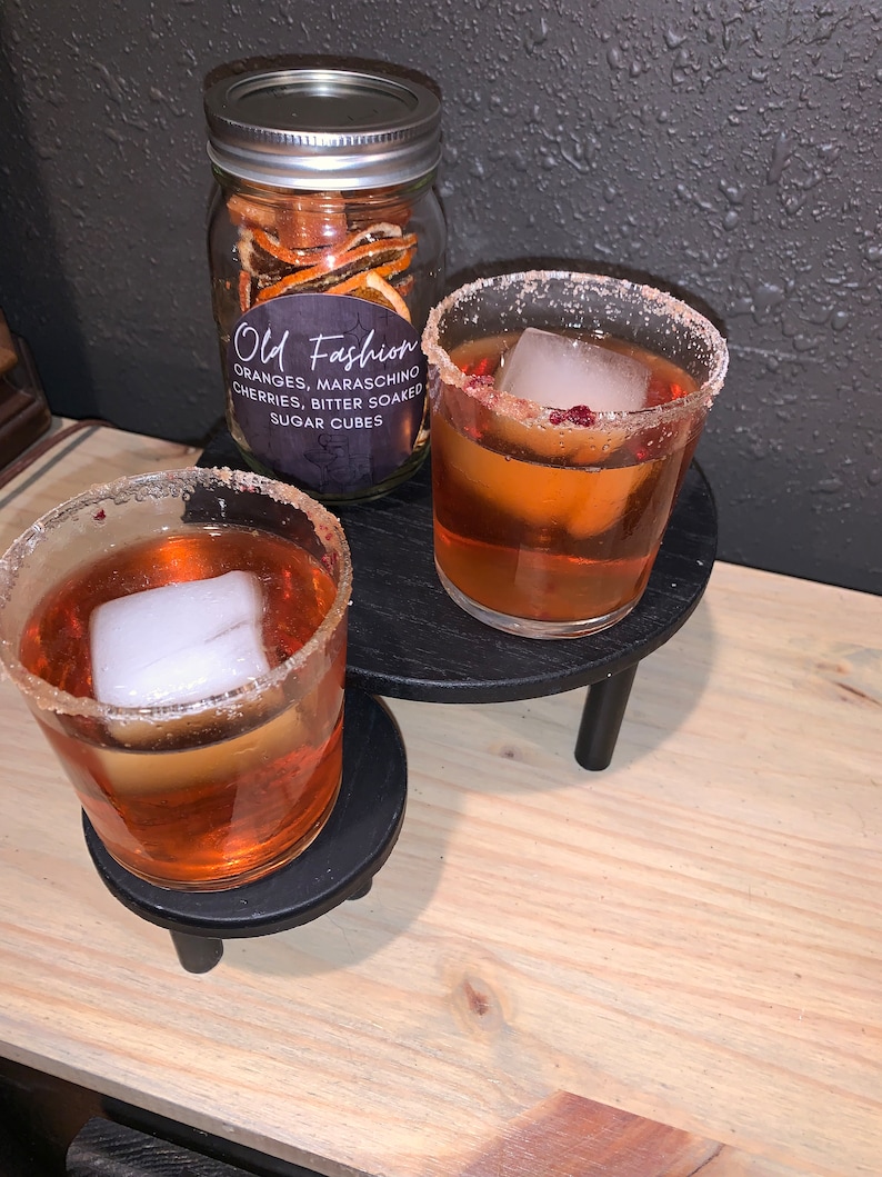 May include: Two clear glasses with orange liquid and ice cubes, rimmed with sugar. The glasses are on black coasters with three legs. A glass jar with orange peels and a label that reads "Old Fashioned Oranges, Maraschino Cherries, Bitter Soaked Sugar Cubes" is in the background.