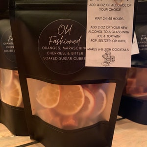 May include: A black pouch with a clear window showing orange and maraschino cherry slices. The pouch has a white tag attached with the text "Old Fashioned" and instructions for making cocktails. The instructions include adding 14 ounces of alcohol of choice, waiting 24-48 hours, adding 2 ounces of new alcohol to a glass with ice and topping with pop, seltzer, or juice. It also states that the pouch makes 6-8 lush cocktails.