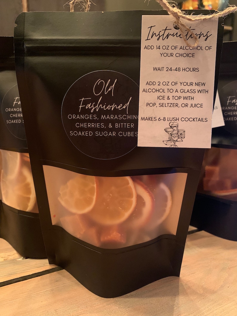 May include: A black pouch with a clear window showing orange and maraschino cherry slices. The pouch has a white tag attached with the text "Old Fashioned" and instructions for making cocktails. The instructions include adding 14 ounces of alcohol of choice, waiting 24-48 hours, adding 2 ounces of new alcohol to a glass with ice and topping with pop, seltzer, or juice. It also states that the pouch makes 6-8 lush cocktails.