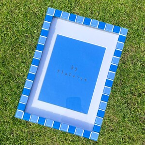 May include: A rectangular picture frame featuring a border of alternating light and dark blue square glass tiles. The frame contains a white mat board surrounding a bright blue centre panel with the text "by florence" printed in small dark letters.