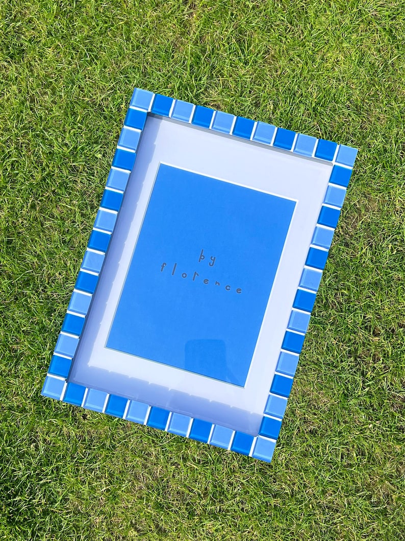 May include: A rectangular picture frame featuring a border of alternating light and dark blue square glass tiles. The frame contains a white mat board surrounding a bright blue centre panel with the text "by florence" printed in small dark letters.