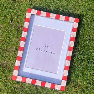 May include: A rectangular picture frame featuring a border of alternating red and light pink square tiles. The frame contains a light purple insert with the text "by florence" printed in a simple, dark, lowercase sans-serif font centred.