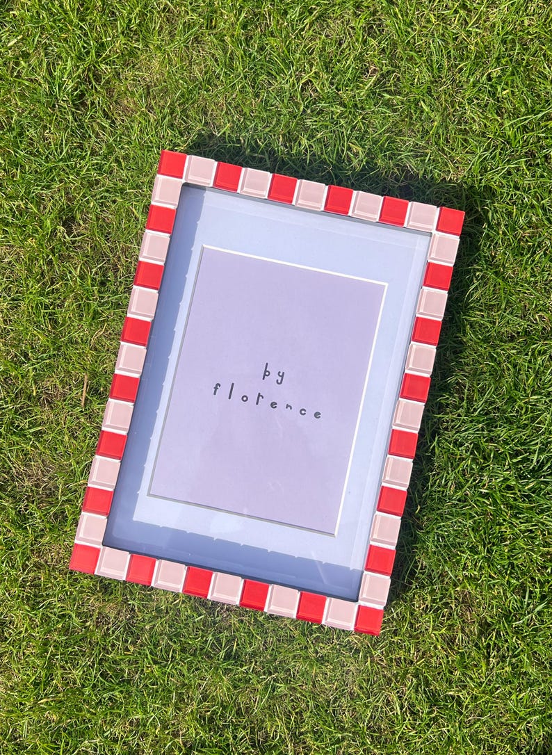 May include: A rectangular picture frame featuring a border of alternating red and light pink square tiles. The frame contains a light purple insert with the text "by florence" printed in a simple, dark, lowercase sans-serif font centred.