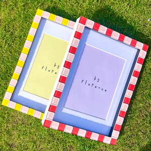 May include: Two rectangular picture frames with mosaic tile borders. The left frame has yellow and light pink tiles with a pale yellow insert reading 'by flore'. The right frame has red and light pink tiles with a purple insert reading 'by florence'.