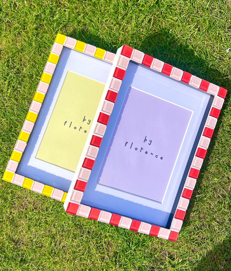 May include: Two rectangular picture frames with mosaic tile borders. The left frame has yellow and light pink tiles with a pale yellow insert reading 'by flore'. The right frame has red and light pink tiles with a purple insert reading 'by florence'.