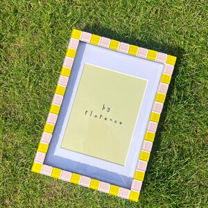 May include: A rectangular picture frame featuring a border of alternating light pink and bright yellow square tiles. The frame contains a white mat and a light green insert with the text "by florence" printed in black, lowercase letters.