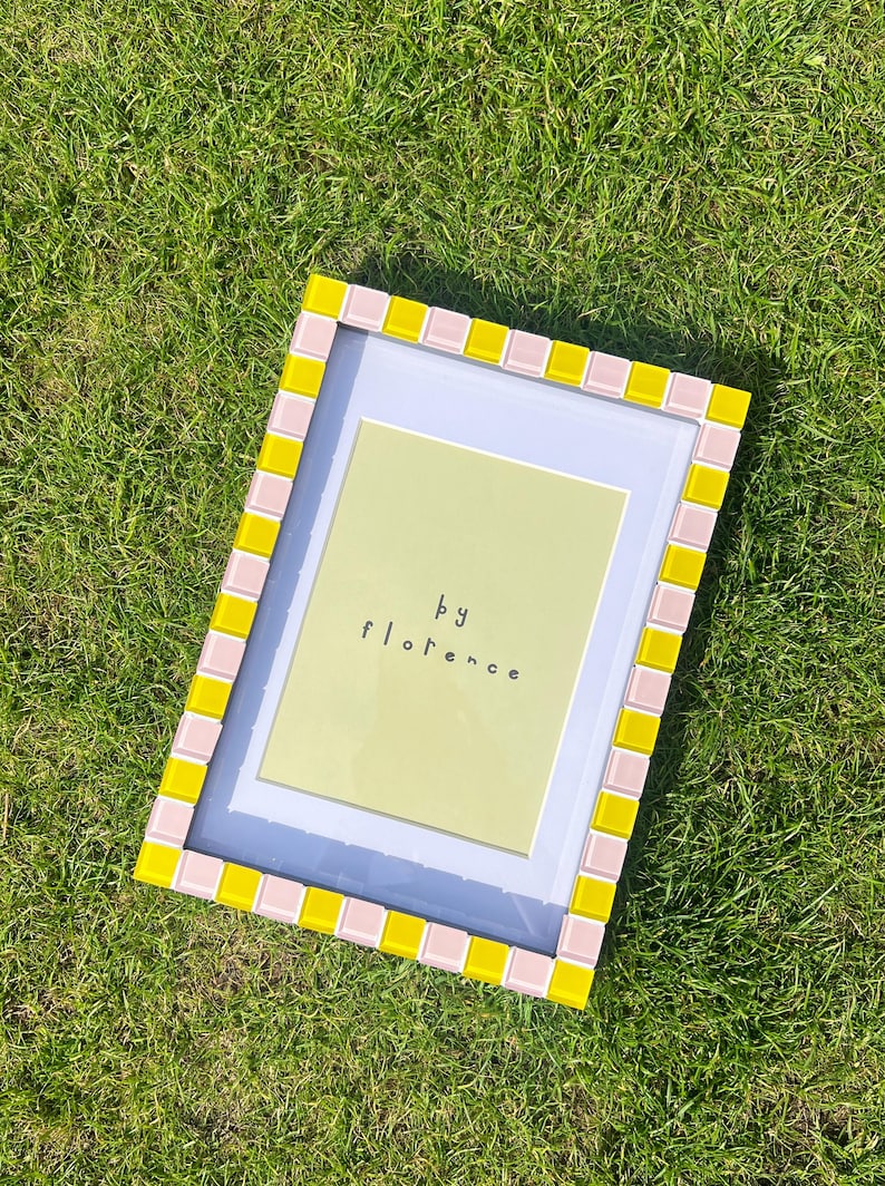 May include: A rectangular picture frame featuring a border of alternating light pink and bright yellow square tiles. The frame contains a white mat and a light green insert with the text "by florence" printed in black, lowercase letters.