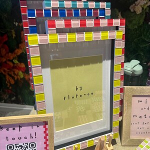 May include: A stack of rectangular picture frames with mosaic tile borders in red, blue, yellow, and pink. The front frame features a yellow insert with the text 'by florence'. Additional frames and signs with text and QR codes are visible nearby.