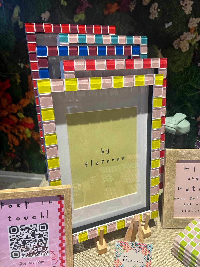 May include: A stack of rectangular picture frames with mosaic tile borders in red, blue, yellow, and pink. The front frame features a yellow insert with the text 'by florence'. Additional frames and signs with text and QR codes are visible nearby.