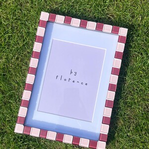 May include: A rectangular picture frame featuring a mosaic border of alternating light pink and deep burgundy square tiles. The frame contains a light purple insert with the text "by florence" printed in a simple, dark, lowercase font.