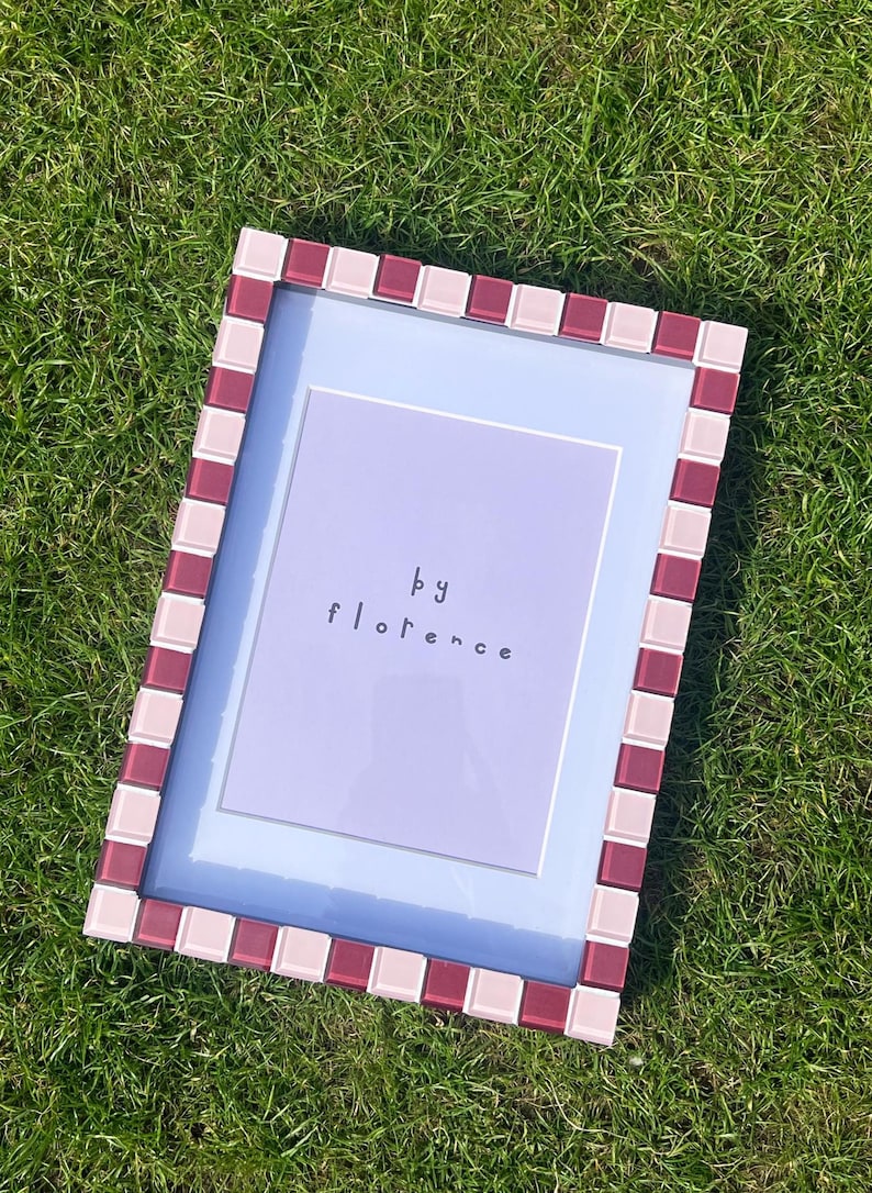 May include: A rectangular picture frame featuring a mosaic border of alternating light pink and deep burgundy square tiles. The frame contains a light purple insert with the text "by florence" printed in a simple, dark, lowercase font.