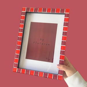 May include: A rectangular picture frame featuring a border of alternating red and dark red glass tiles. The frame contains a white mat surrounding a dark red insert with the text "by florence" printed in black in the centre of the display area.