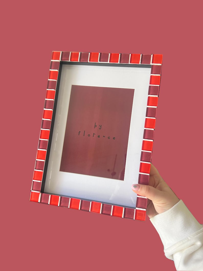 May include: A rectangular picture frame featuring a border of alternating red and dark red glass tiles. The frame contains a white mat surrounding a dark red insert with the text "by florence" printed in black in the centre of the display area.